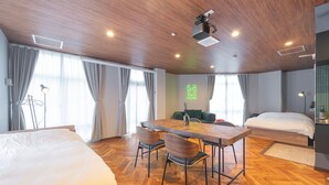 A hotel room with a wooden ceiling, a dining table with chairs, a bed, and a large window with curtains.