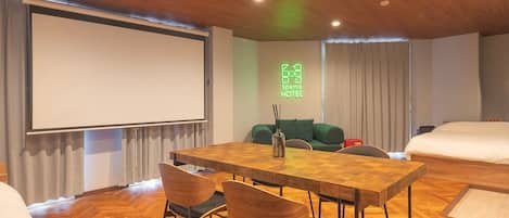 A hotel room with a wooden ceiling, a large bed, a sofa, a dining table with chairs, and a projector screen.