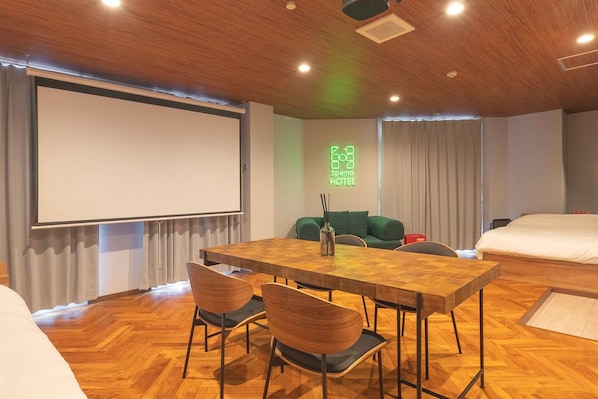 A hotel room with a wooden ceiling, a large bed, a sofa, a dining table with chairs, and a projector screen.