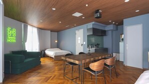 A hotel room with a wooden ceiling, a bed, a green sofa, a dining table with chairs, and a kitchenette.