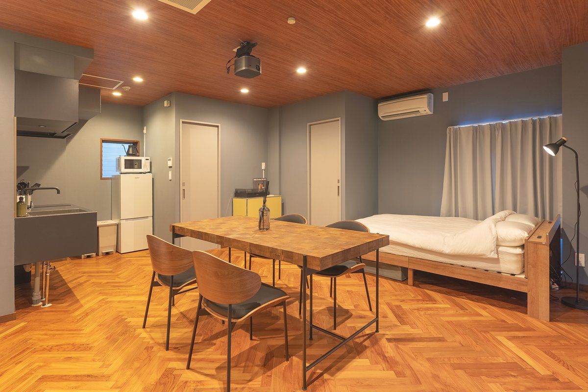 Ein kompakter Raum mit Holzboden, einem Bett, einem Schreibtisch mit Stuhl, einer kleinen Küchenzeile und einem Deckenprojektor.