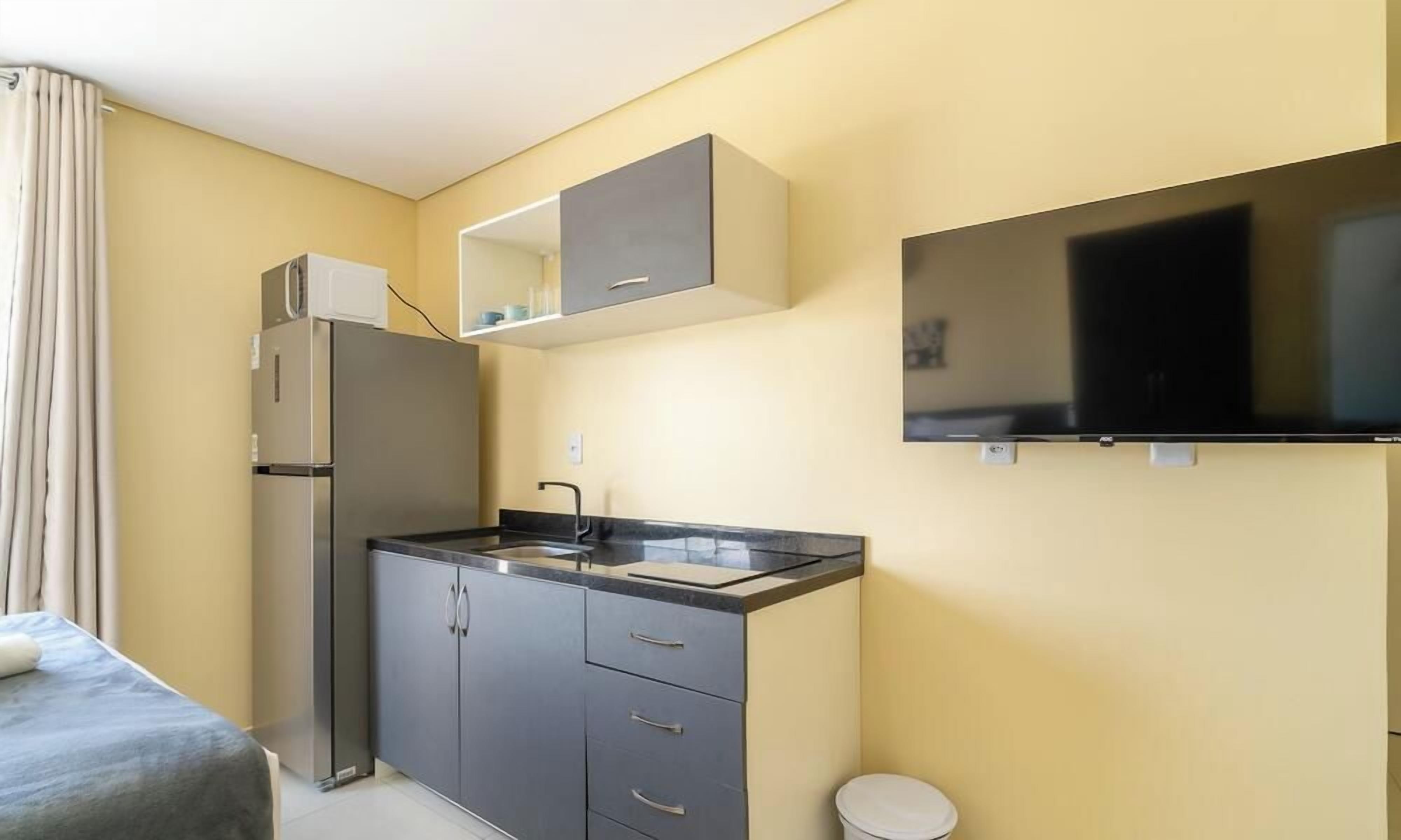 Una kitchenette compacta con refrigerador, pileta y placares, un televisor de pantalla plana montado en la pared, y una cama individual con cortina.
