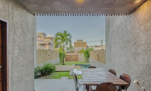 Um alpendre coberto com mesa e cadeiras de madeira, uma pequena piscina e vista para uma área residencial com casas e palmeiras.