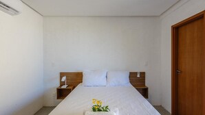 Una cama bien hecha con edredón blanco, cabecera de madera y mesita de noche con lámpara.