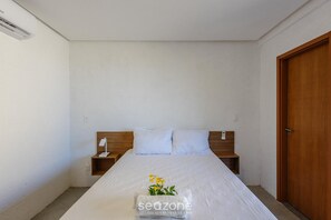 Una cama bien hecha con edredón blanco, cabecera de madera y mesita de noche con lámpara.