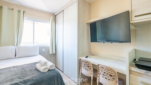 Quarto de hotel com cama, escrivaninha com duas cadeiras, televisão de tela plana e janela com cortinas.