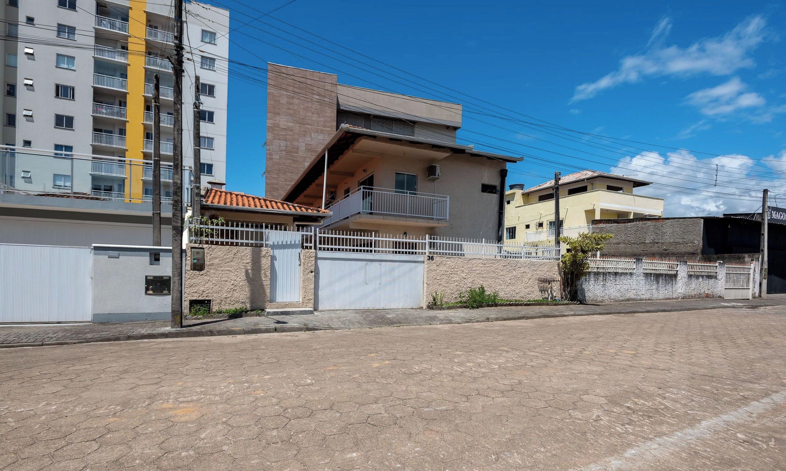 A residential street with a two-story house featuring a balcony, a white fence, and a paved road.