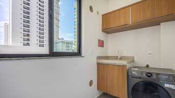 A compact laundry room with a washing machine, wooden cabinets, a window with a view of buildings, and a small sink.