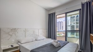 Ett hotellrum med en sänggavel i marmor, en säng med vita sängkläder, ett grått överkast och utsikt över byggnader genom ett stort fönster.