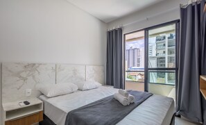 Quarto de hotel com cabeceira de mármore, cama com roupa de cama branca, manta cinzenta e vista para edifícios através de uma grande janela.