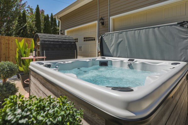 Un jacuzzi en un patio trasero con valla de madera, plantas en macetas y un garaje al fondo.