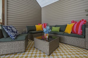 Un área de patio techada con muebles de mimbre, incluyendo un sofá y dos sillones, una mesa de centro y un jarrón decorativo con flores.