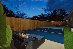 Un court de tennis en plein air, entouré d’une clôture en bois, agrémenté de guirlandes lumineuses et doté d’un espace de repos couvert avec des parasols.