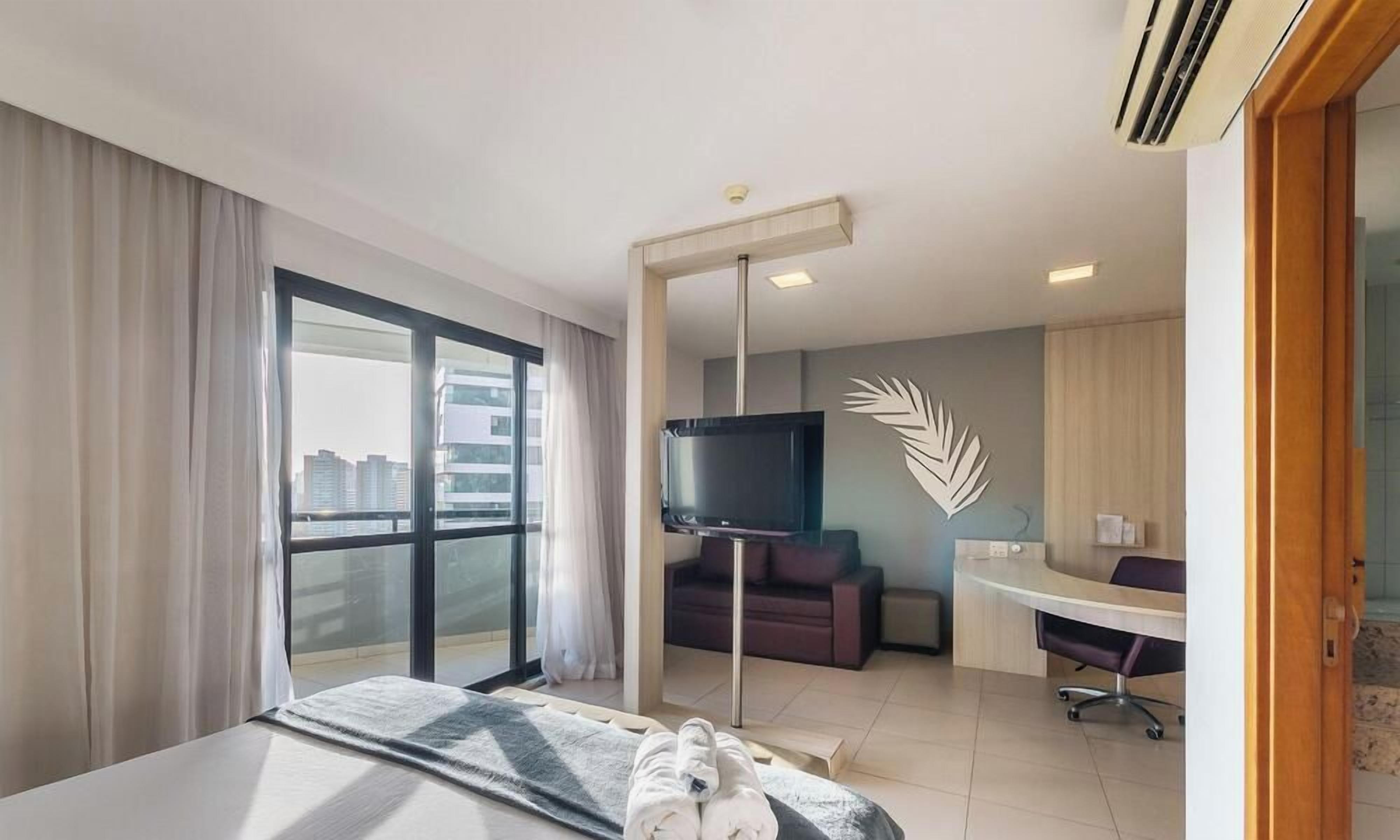 A hotel room with a bed, a desk, a TV, a sofa, and a large window with a city view.