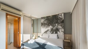 Moderni hotellihuone, jossa on sänky, yöpöytä, seinälle ripustettu taideteos, suihkutila ja ovi, joka johtaa toiseen huoneeseen.