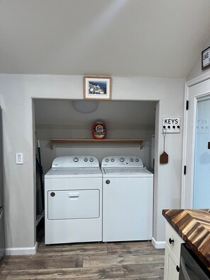Une buanderie équipée de deux machines à laver et de deux sèche-linge superposés, d’une étagère sur laquelle se trouve une boîte de lessive Tide, et d’une porte sur laquelle est affiché un panneau indiquant « CLÉS ».