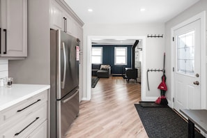 A modern kitchen with a stainless steel refrigerator, wooden flooring, and a view into a living area with a sofa and a fireplace.