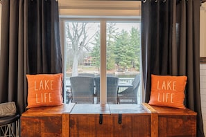A room with a view of a lake, featuring two orange pillows with 'LAKE LIFE' text, a wooden bench, and wicker chairs.