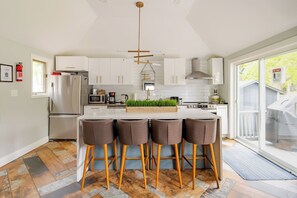 A modern kitchen with white cabinets, a central island with bar stools, a stainless steel refrigerator, and a large window.