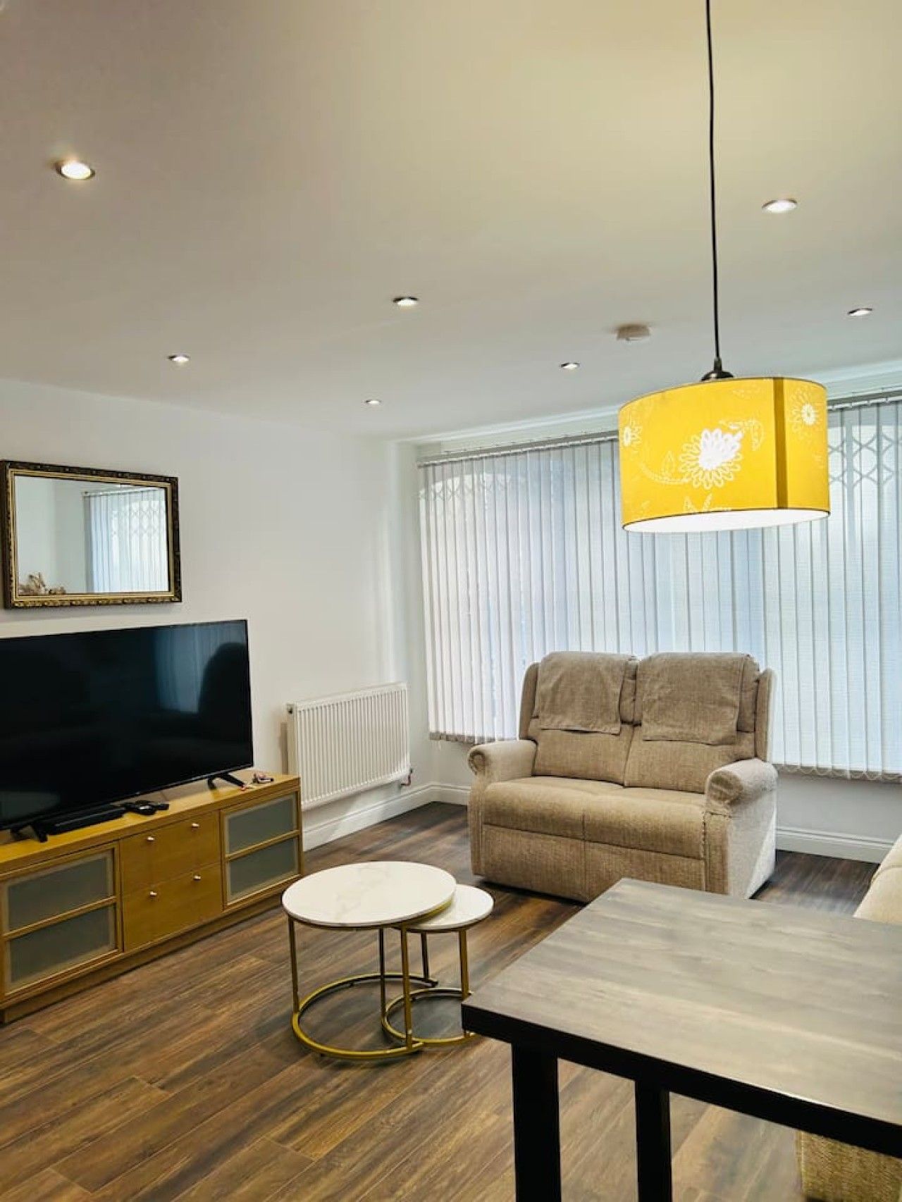 A modern living room with a flat-screen TV, a beige sofa, a round white table, and a yellow pendant light.