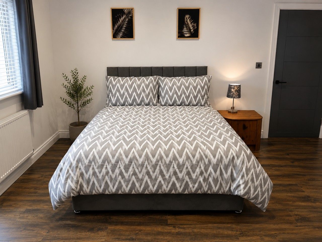 A bedroom with a bed, a nightstand, a lamp, a potted plant, and two framed pictures on the wall.