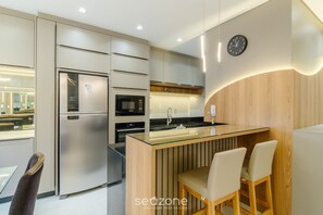 Une cuisine moderne équipée d’un réfrigérateur, d’un four à micro-ondes et d’une cuisinière intégrés. On y trouve un îlot central en verre avec des sièges et une horloge murale.