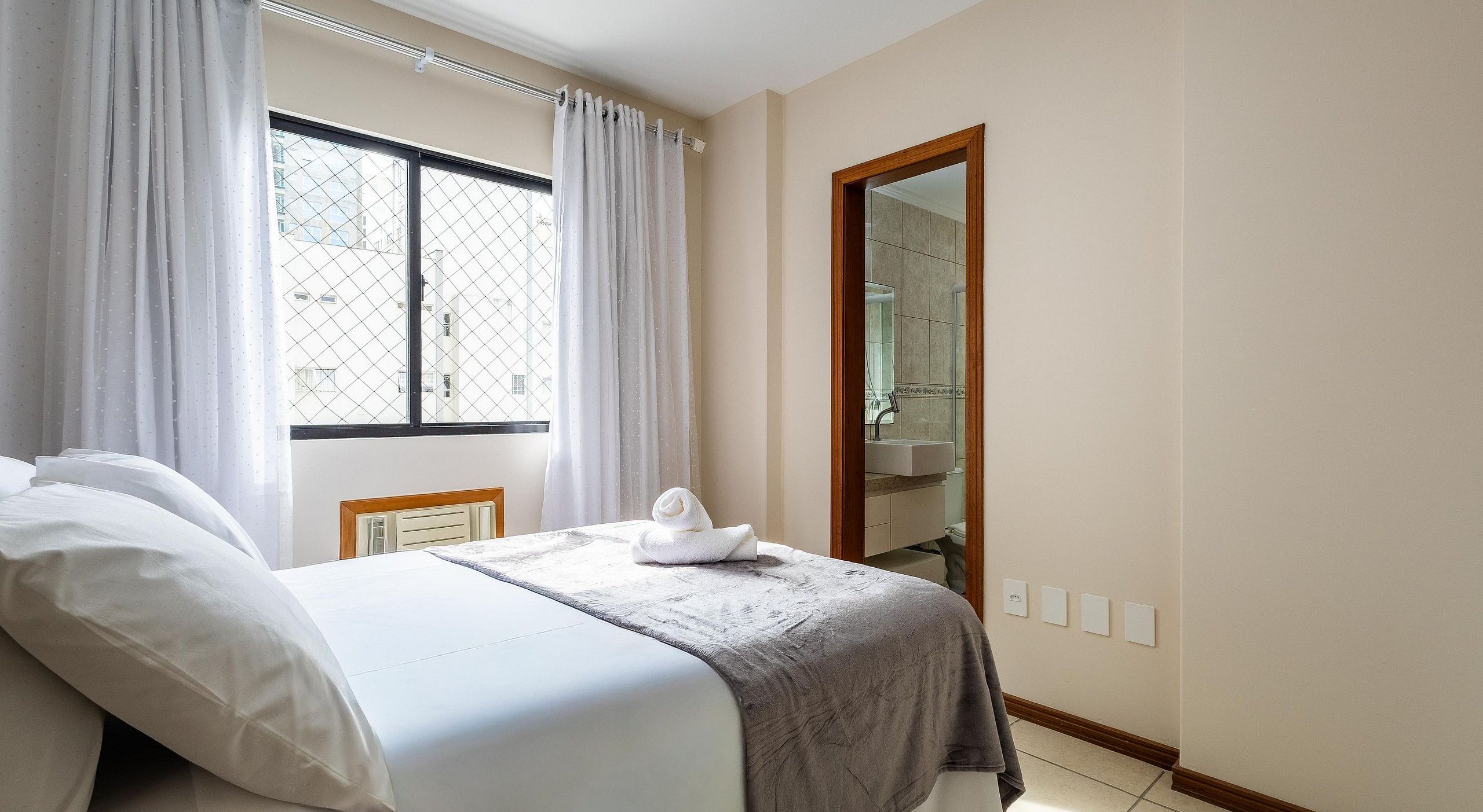 A hotel room with a bed, a window with curtains, a bathroom visible through a mirror, and a tiled floor.