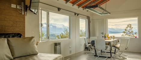 A room with a bed, a dining table with chairs, large windows with mountain views, and a ceiling with wooden beams.