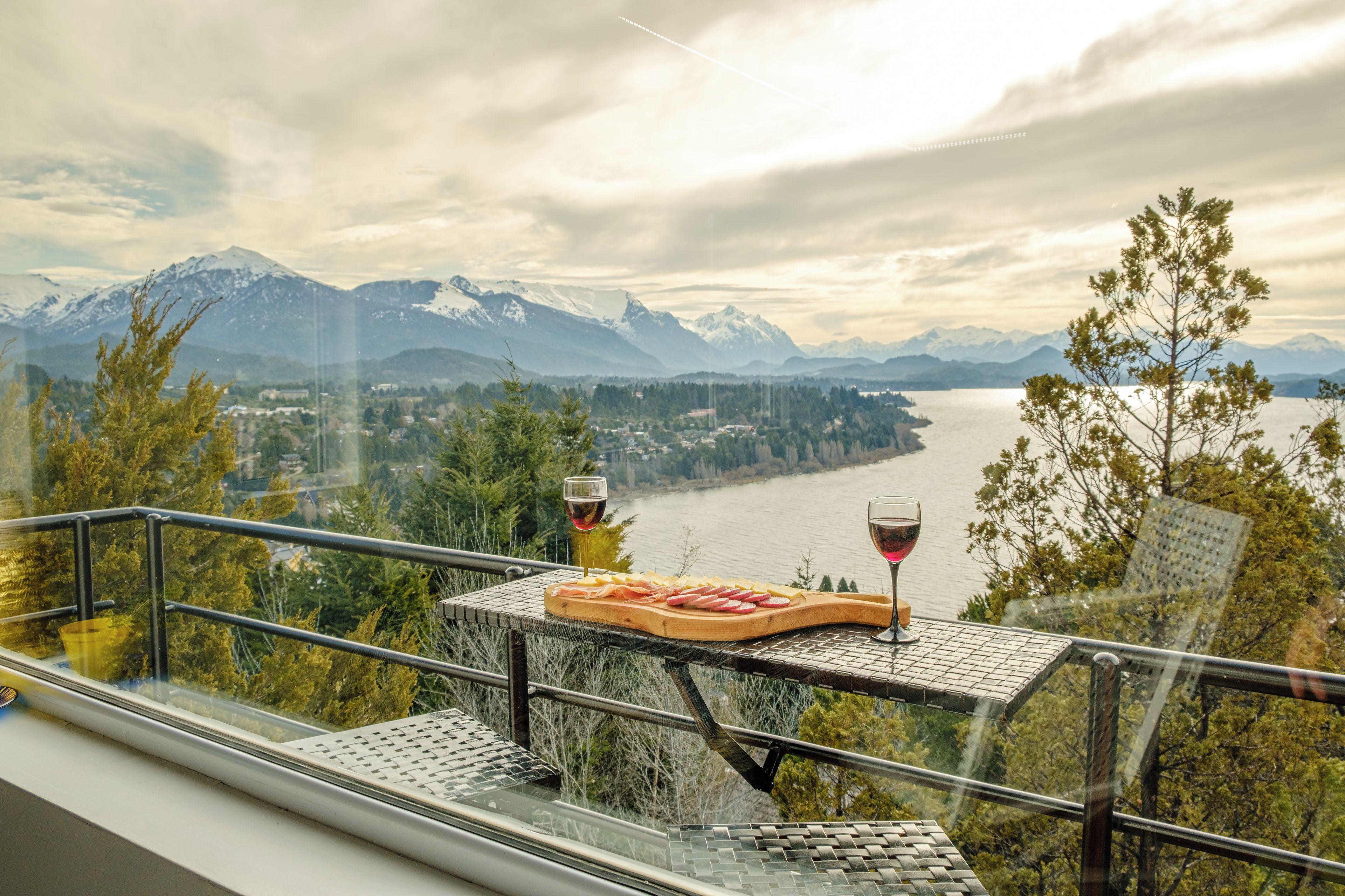 A table with a cheese platter and wine glasses, overlooking a lake and mountains.