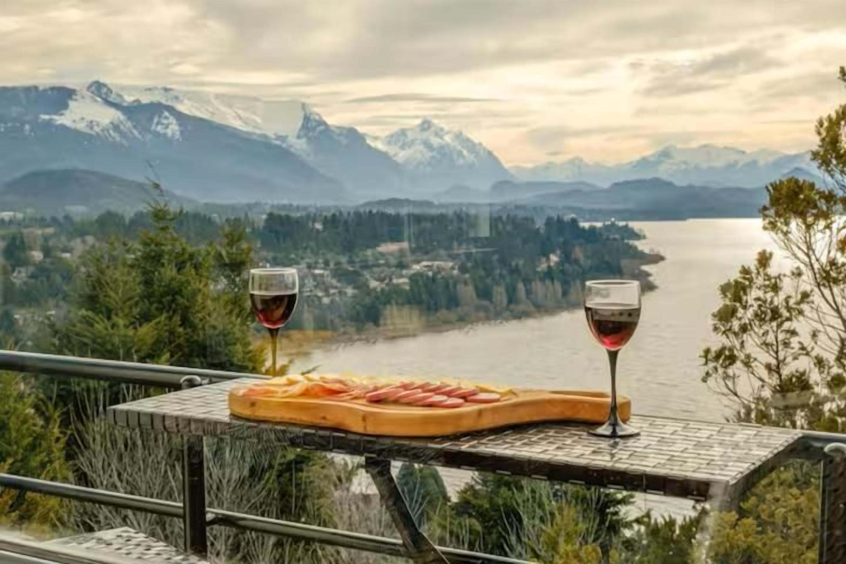 Una mesa con una tabla de quesos y dos copas de vino tinto, con vistas a un lago y montañas.