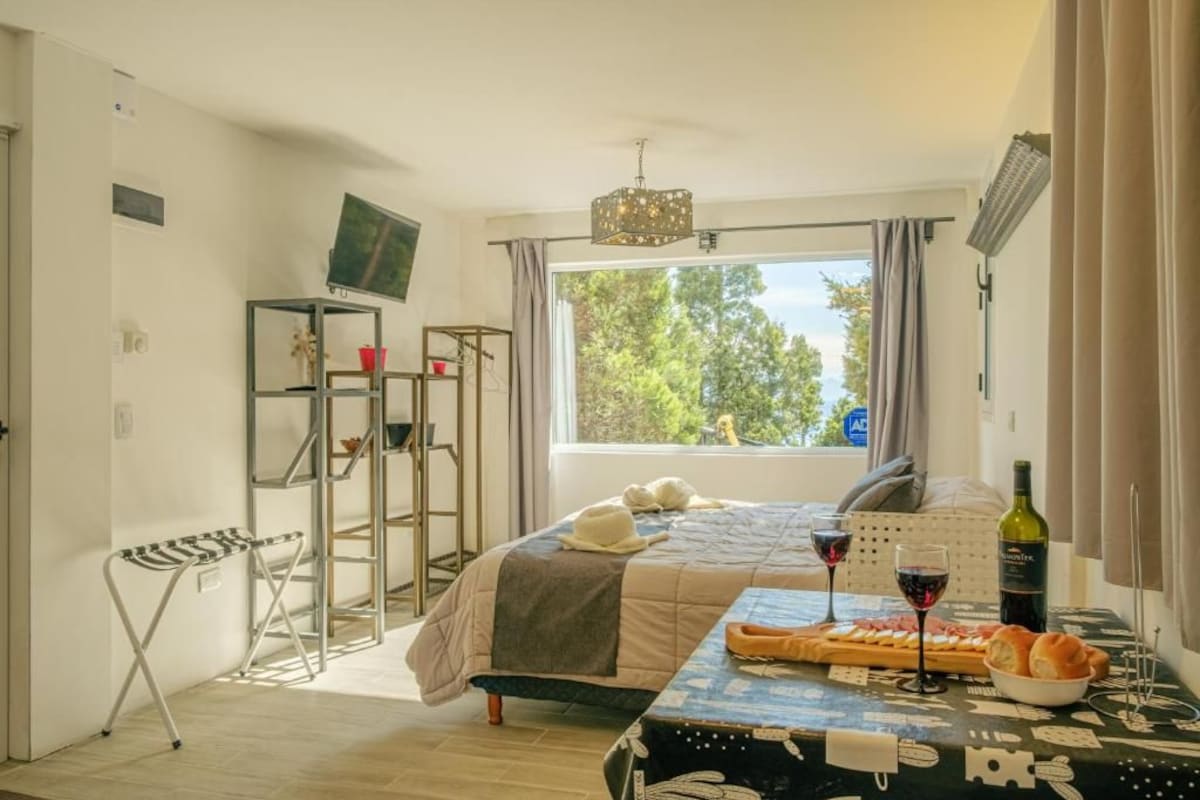 A bedroom with a bed, a table with a bottle and glasses, a chair, a TV on a stand, and a window with a view of trees.
