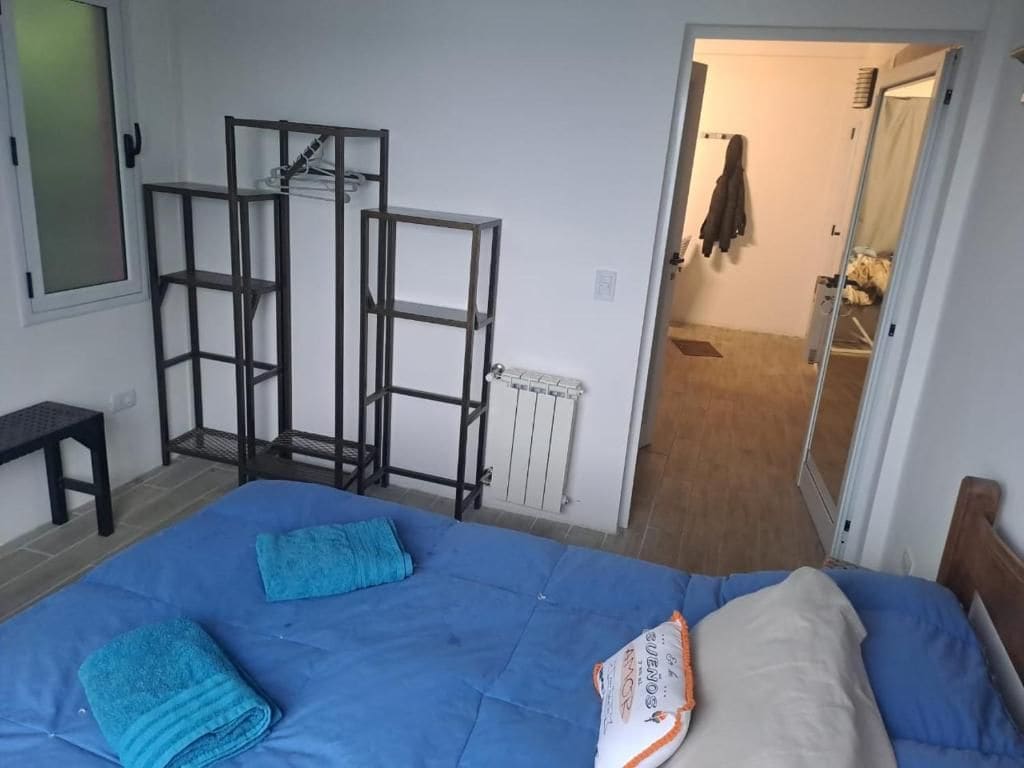 Un dormitorio con cama, colchón azul y almohada blanca. Se observa un perchero metálico contra la pared y una puerta que da a otra habitación.