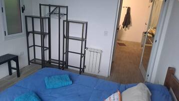 Un dormitorio con cama, colchón azul y almohada blanca. Se observa un perchero metálico contra la pared y una puerta que da a otra habitación.