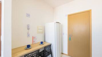 A small kitchenette with a refrigerator, a wooden table with two cups, a chair, and a door with a key lock.