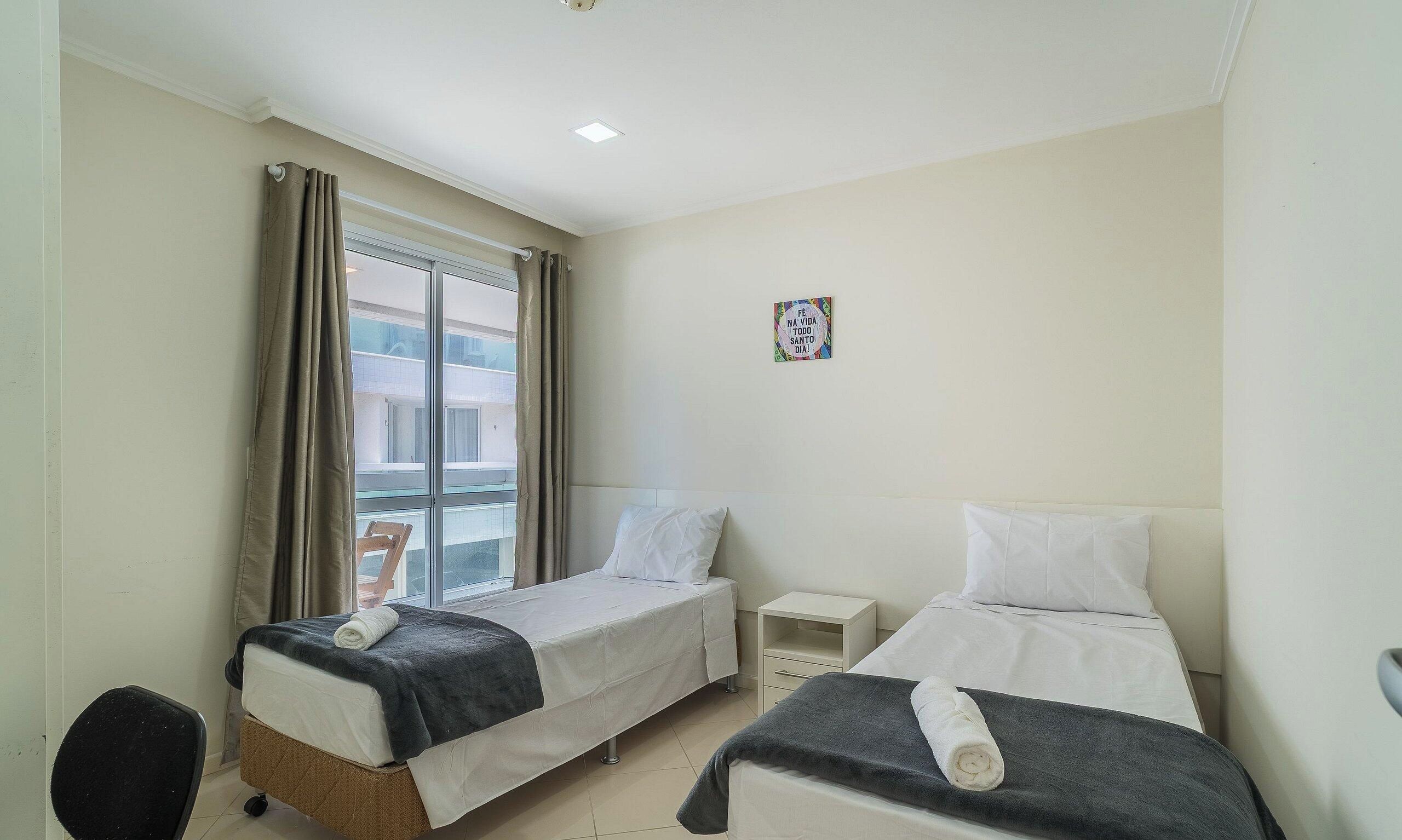 A hotel room with two beds, a small bedside table, a chair, and a window with curtains.
