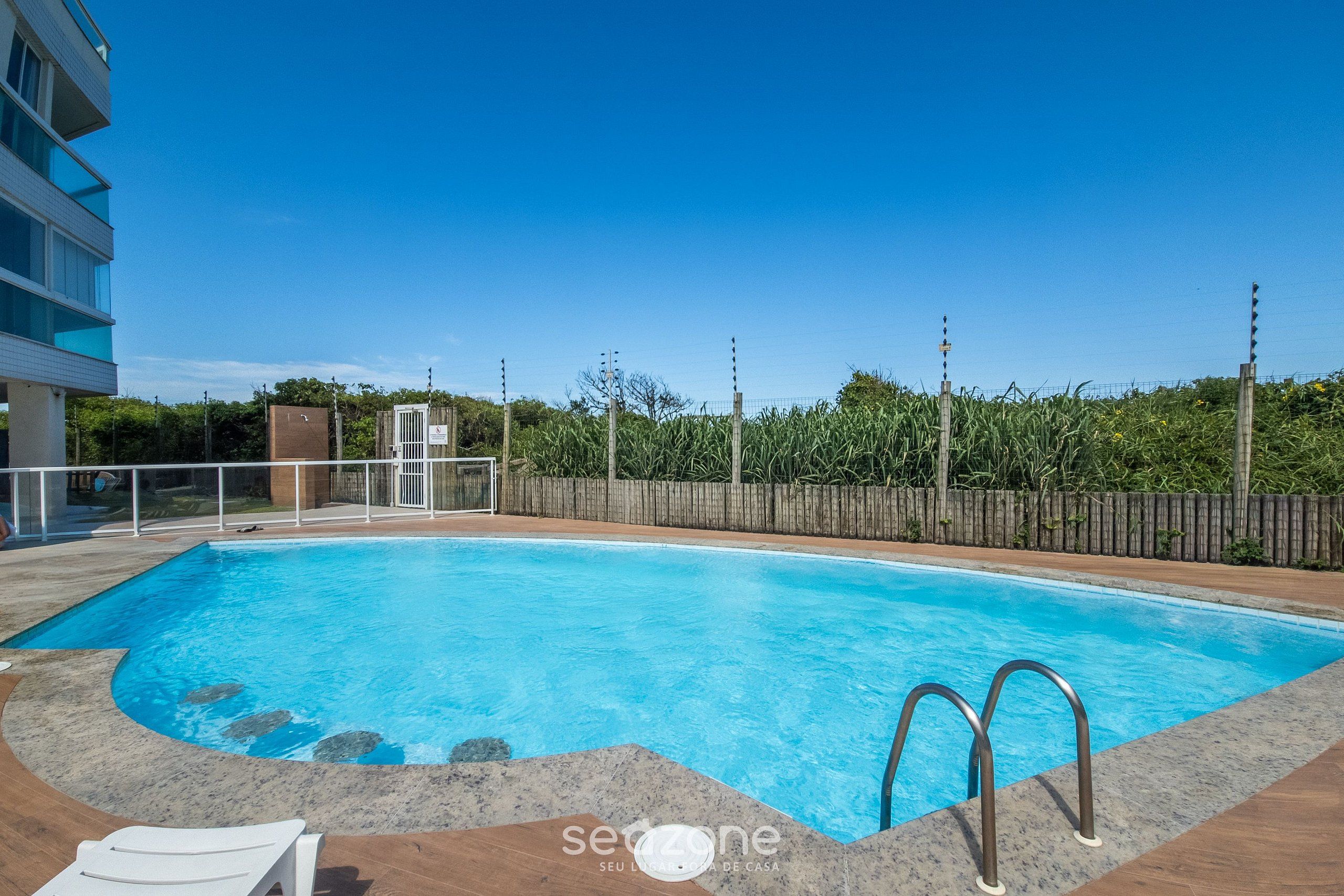 A swimming pool with a wooden deck and metal railings, surrounded by a fence and greenery.