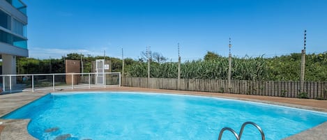 A swimming pool with a wooden deck and metal railings, surrounded by a fence and greenery.