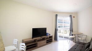 Ein Wohnzimmer mit einem Flachbildfernseher auf einem Holzboden, einer Couch, einem kleinen runden Tisch mit Stühlen und einer Schiebetür zu einem Außenbereich.