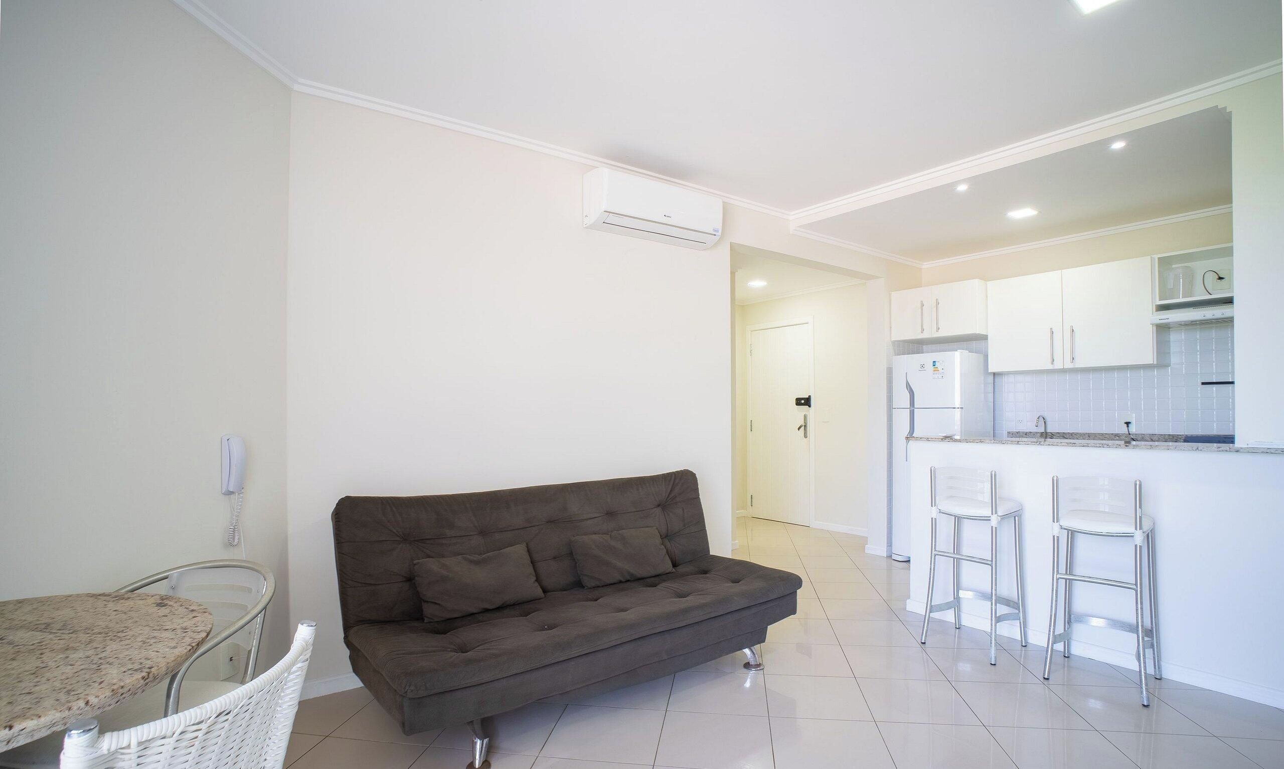 Un appartement moderne avec un canapé gris, une table à manger, une cuisine dotée de meubles blancs et un sol carrelé de couleur blanche.