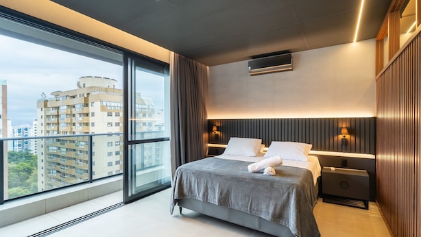 Ett modernt hotellrum med en stor säng, två sängbord, en väggmonterad luftkonditioneringsanläggning och utsikt över staden genom fönster som sträcker sig från golv till tak.