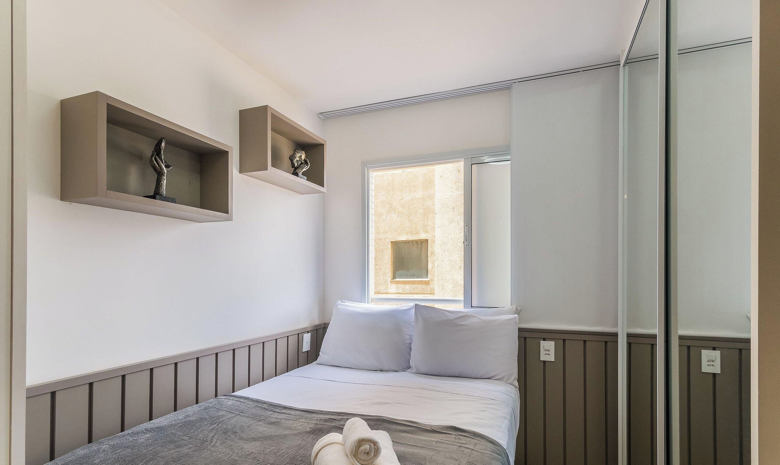 Un dormitorio con una cama, dos estantes en la pared con esculturas, una ventana y un espejo.