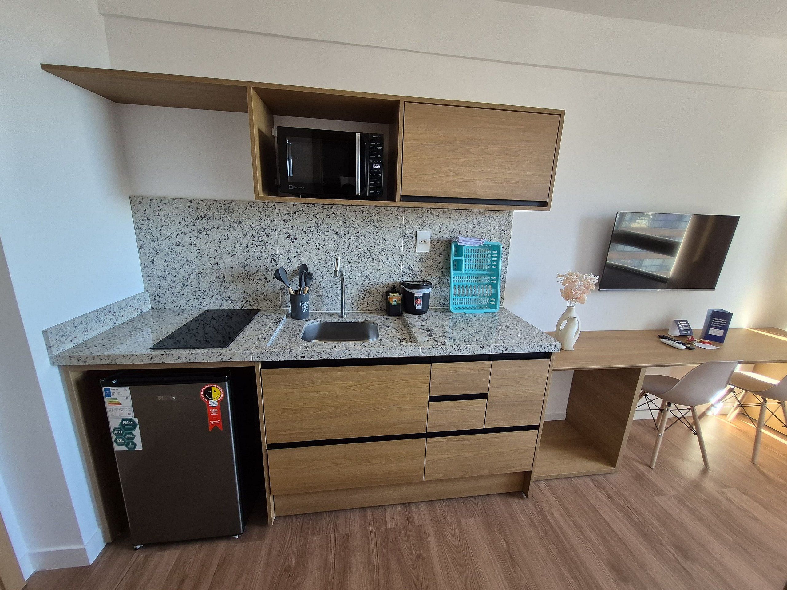 A compact kitchen with a microwave, sink, and a small refrigerator. There is a wooden countertop, a teal dish rack, and a small table with a chair.