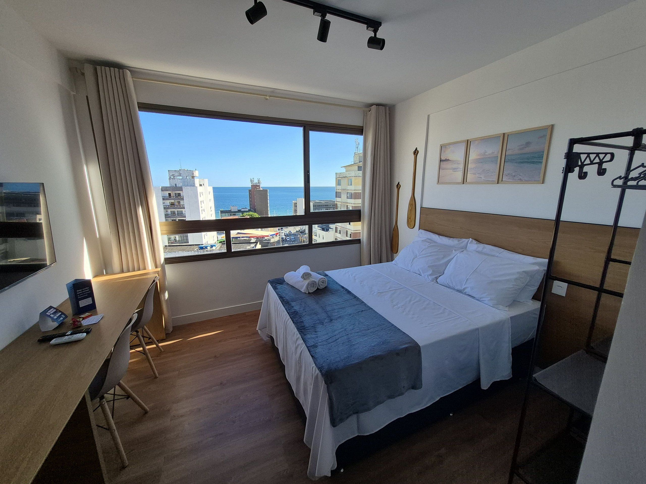 A hotel room with a large window offering a city view, a bed with a blue bedspread, a desk with a chair, and a small table with a chair.