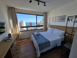 A hotel room with a large window offering a city view, a bed with a blue bedspread, a desk with a chair, and a small table with a chair. - Studio in Build. w/ Pool, 270m fr the sea Cao0903 (Salvador)