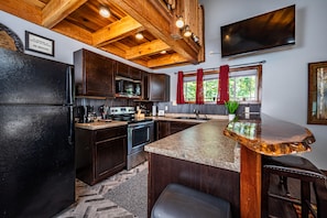 A kitchen with dark wood cabinets, a stainless steel refrigerator, a microwave, and a large island with a granite countertop. - Romantic, Secluded Log Cabin | 2BR, 1.5BA (Columbus)