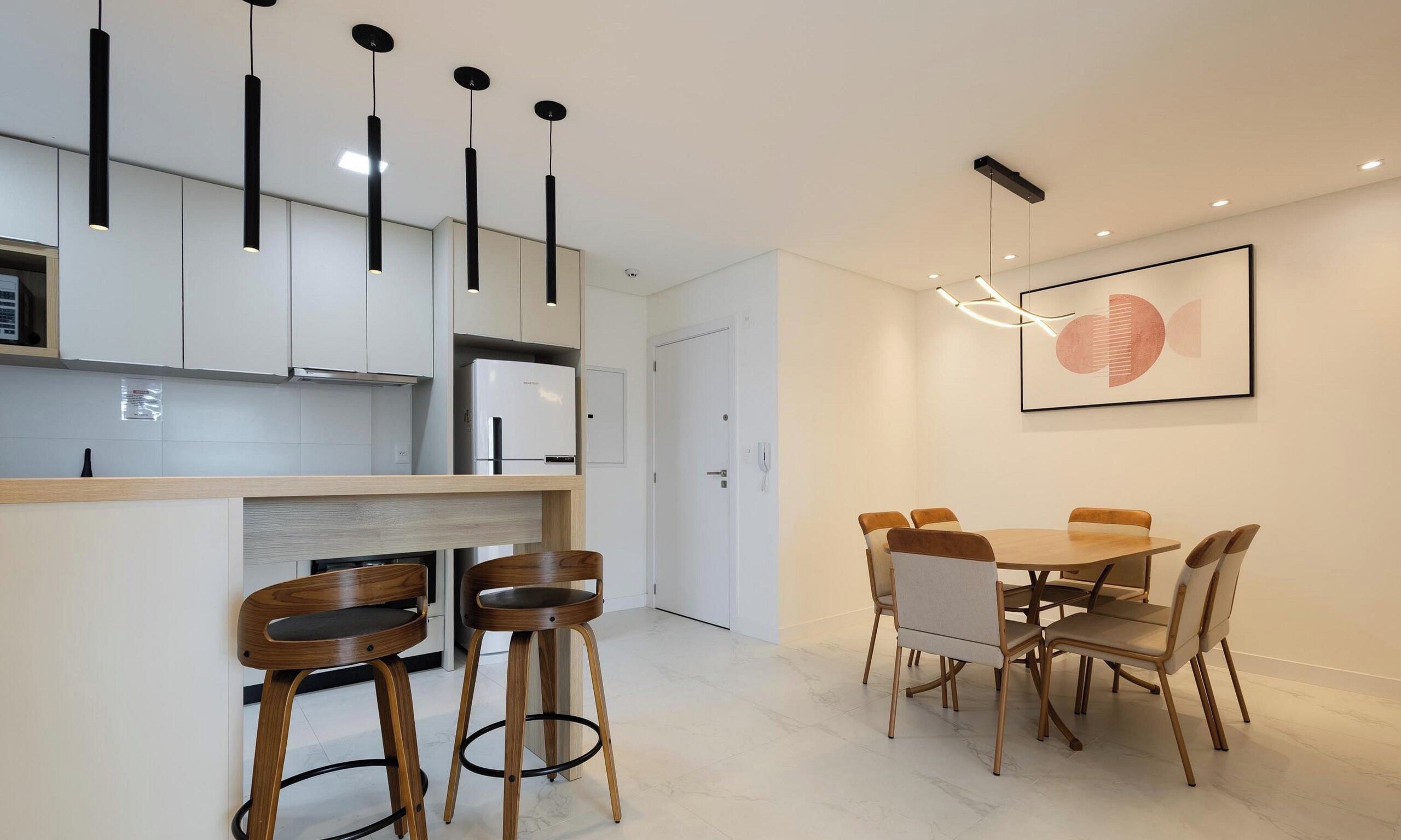 Una cocina moderna con armarios blancos, horno empotrado y refrigerador. Hay un comedor con una mesa y sillas de madera.
