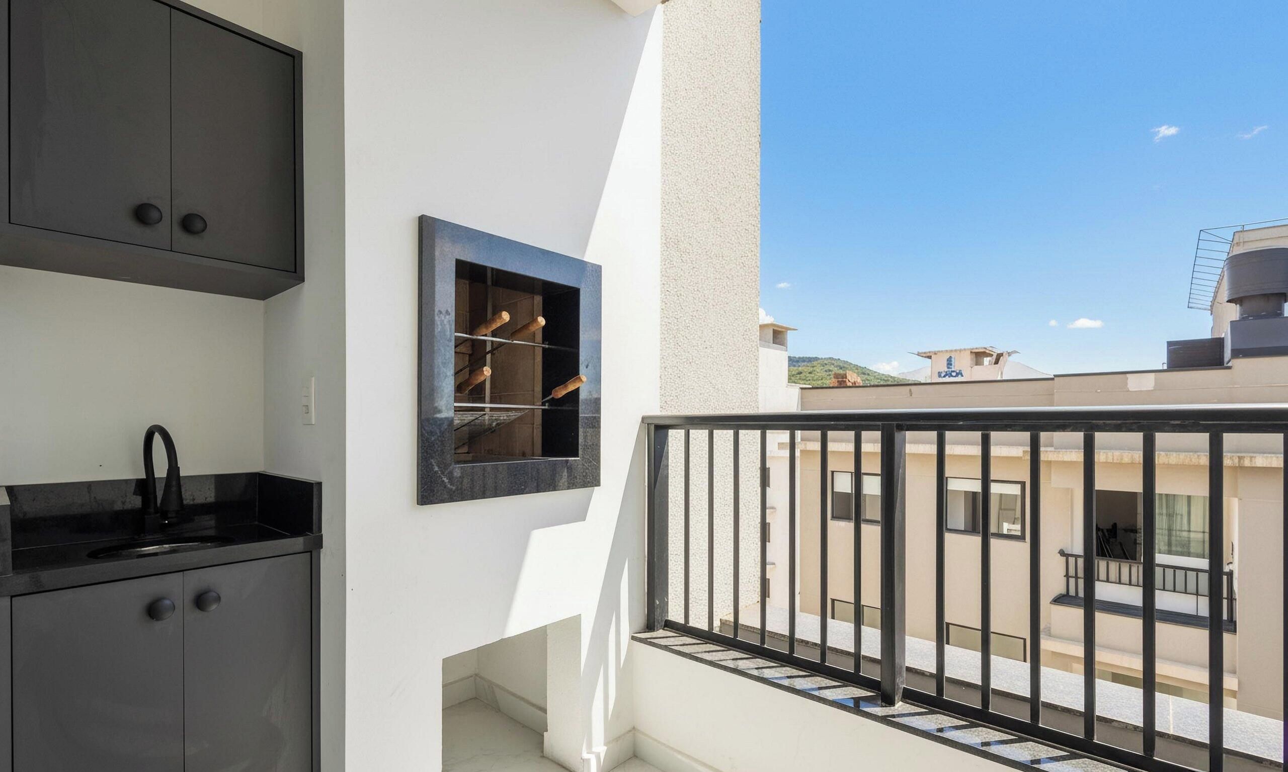 Un balcón con barandilla negra, una pequeña cocina con pileta y muebles negros, y un ventanal con vistas a edificios.
