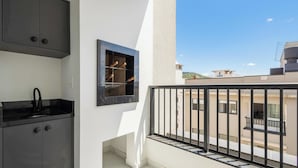 Varanda com guarda-corpo preto, pequena cozinha com pia e armários pretos, e janela com vista para edifícios.