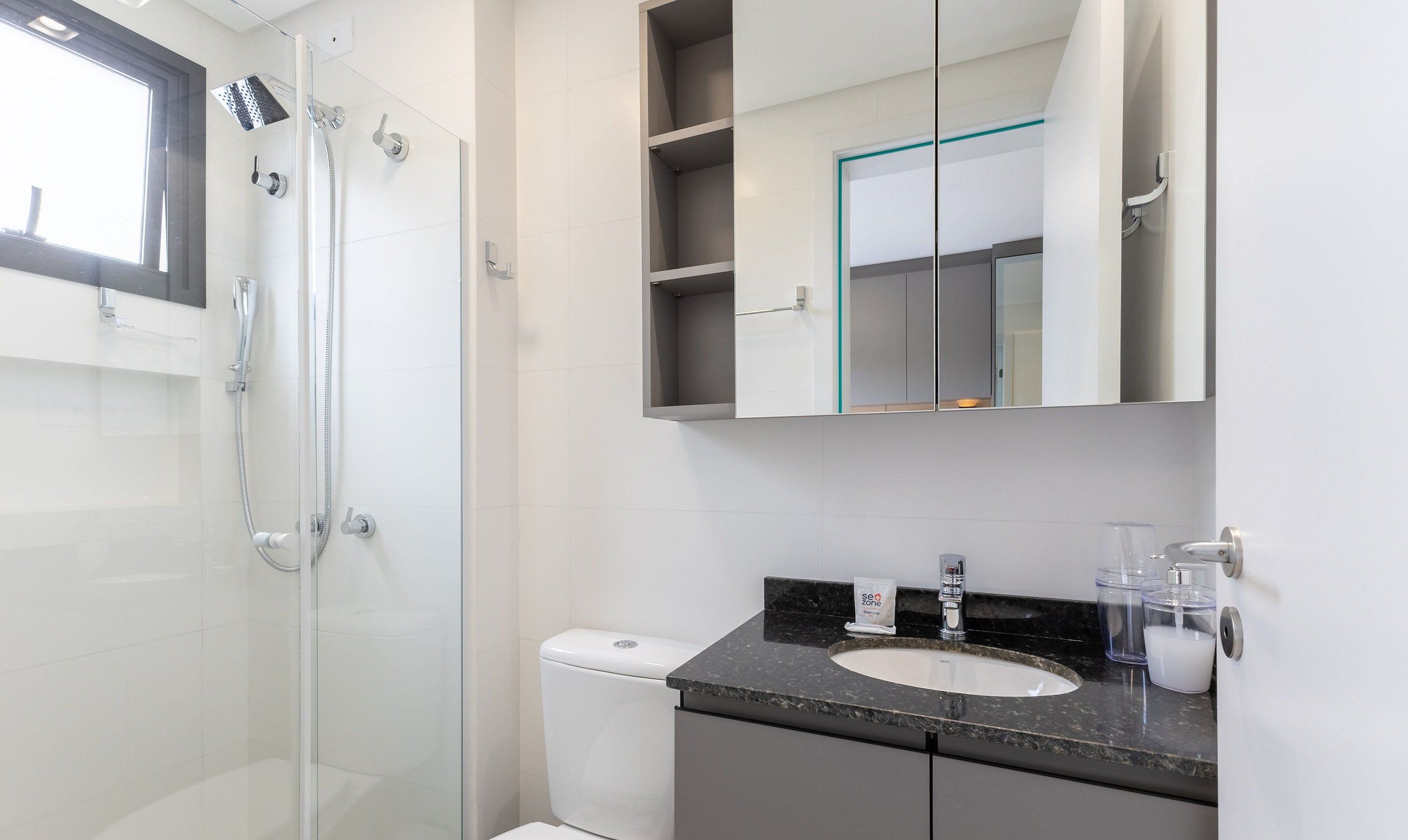 Ein modernes Badezimmer mit Duschkabine aus Glas, Toilette, Waschbecken mit Granitplatte und Spiegel mit Ablagefläche darüber.