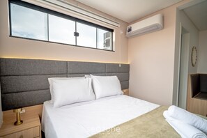 Ein Hotelzimmer mit einem großen Bett, einem Nachttisch, einer Lampe, einem Fenster mit Jalousien, einer Klimaanlage und einem Spiegel.